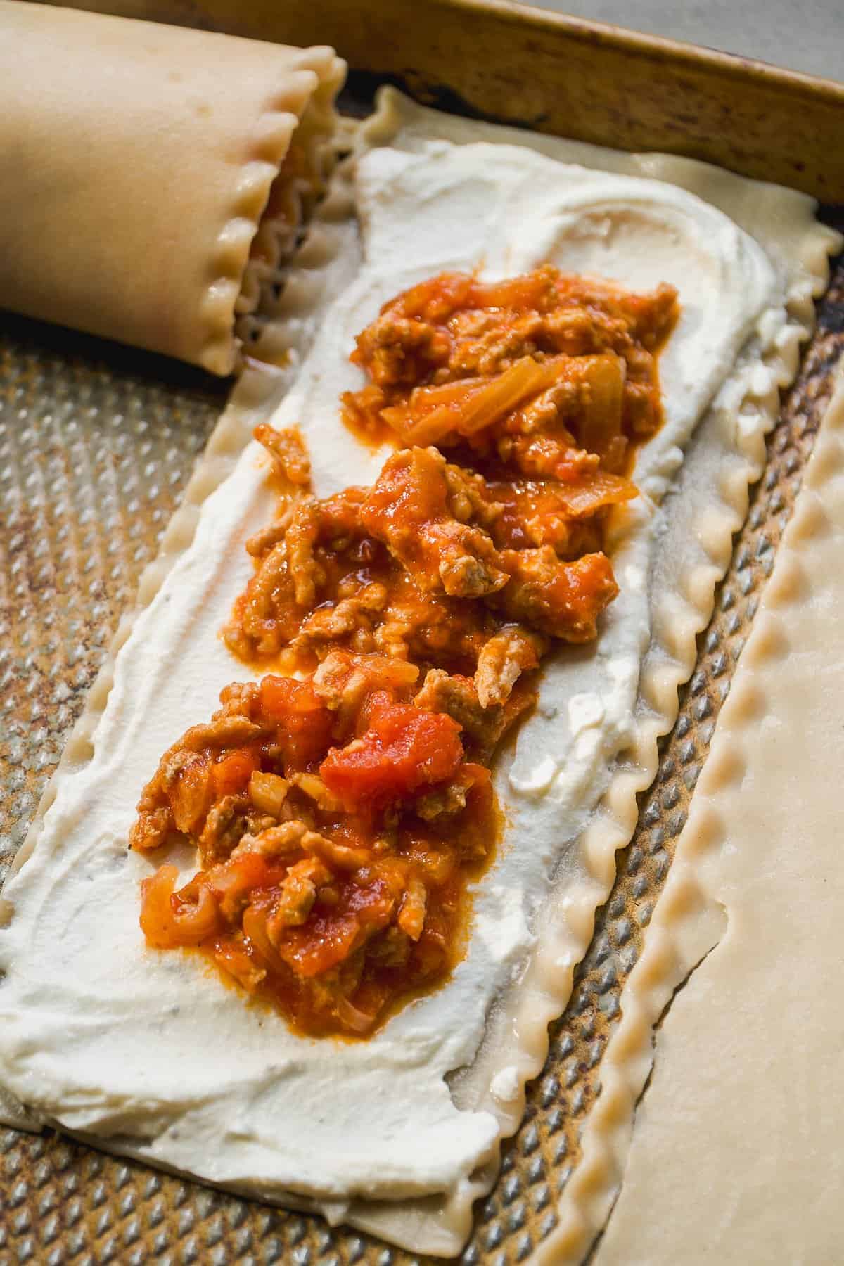 A sheet of pasta is topped with a layer of creamy ricotta cheese and a generous scoop of tomato meat sauce, ready to be rolled or folded, on a textured baking tray.