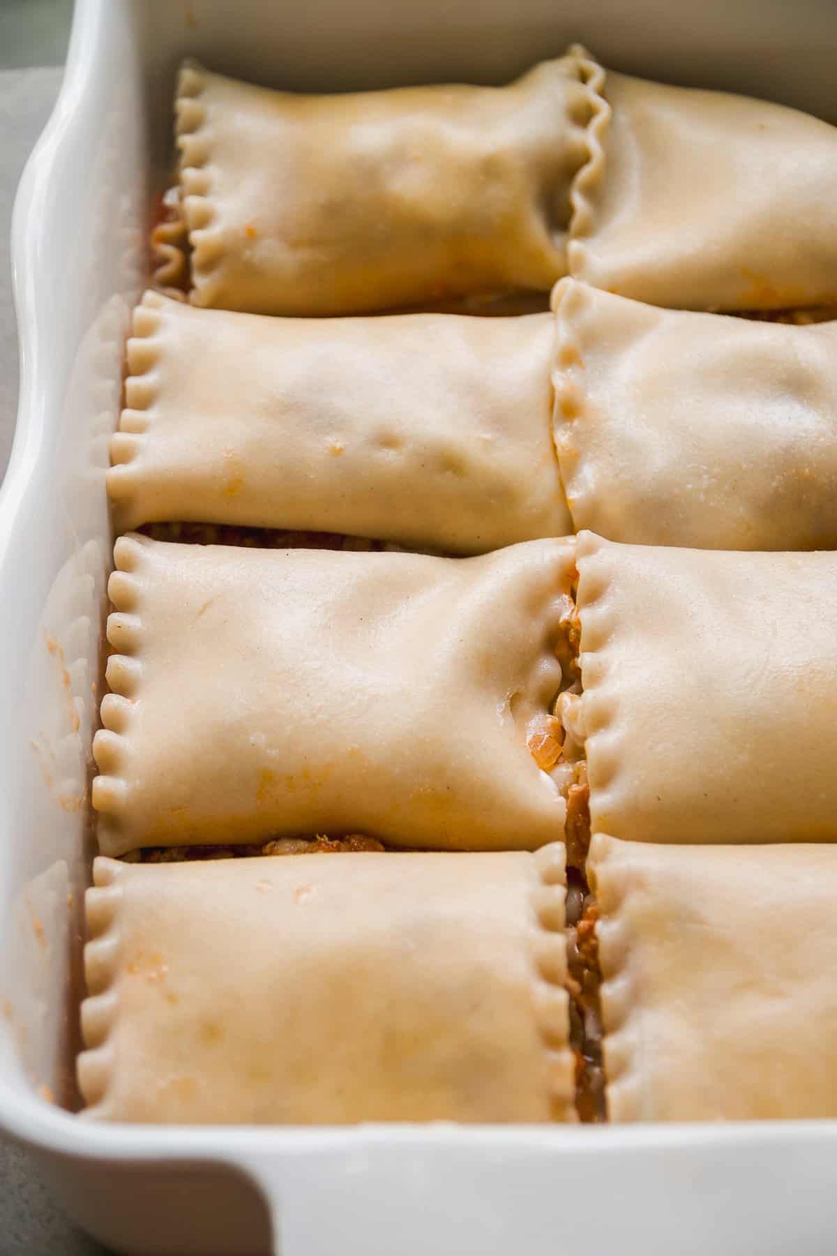 Nine unbaked lasagna roll-ups arranged in a white rectangular baking dish, with crimped edges sealing the pasta and filling inside.
