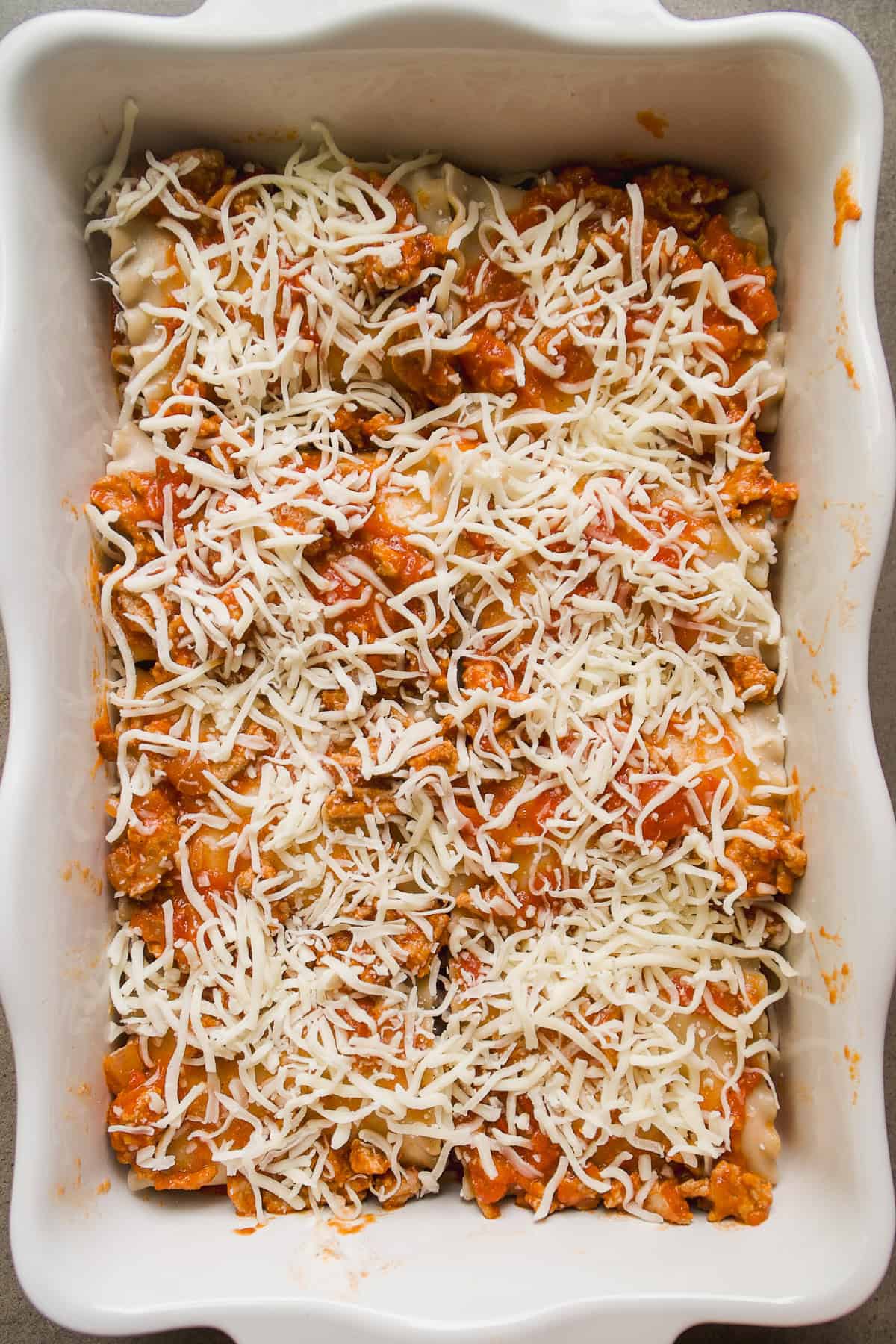A white baking dish filled with unbaked lasagna layers, topped with meat sauce and shredded mozzarella cheese, ready to be cooked.