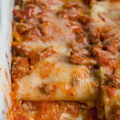 A close-up of a baked lasagna in a white dish, showing layers of pasta, melted cheese, tomato sauce, and browned ground meat.