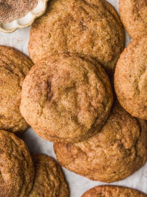 A close-up of several golden-brown snickerdoodle cookies, sprinkled with cinnamon sugar, arranged on a light surface with a small dish of extra cinnamon sugar in the corner.