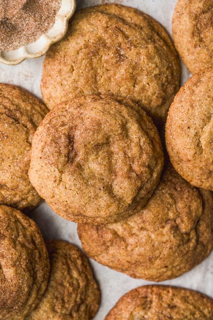 A close-up of several golden-brown snickerdoodle cookies, sprinkled with cinnamon sugar, arranged on a light surface with a small dish of extra cinnamon sugar in the corner.
