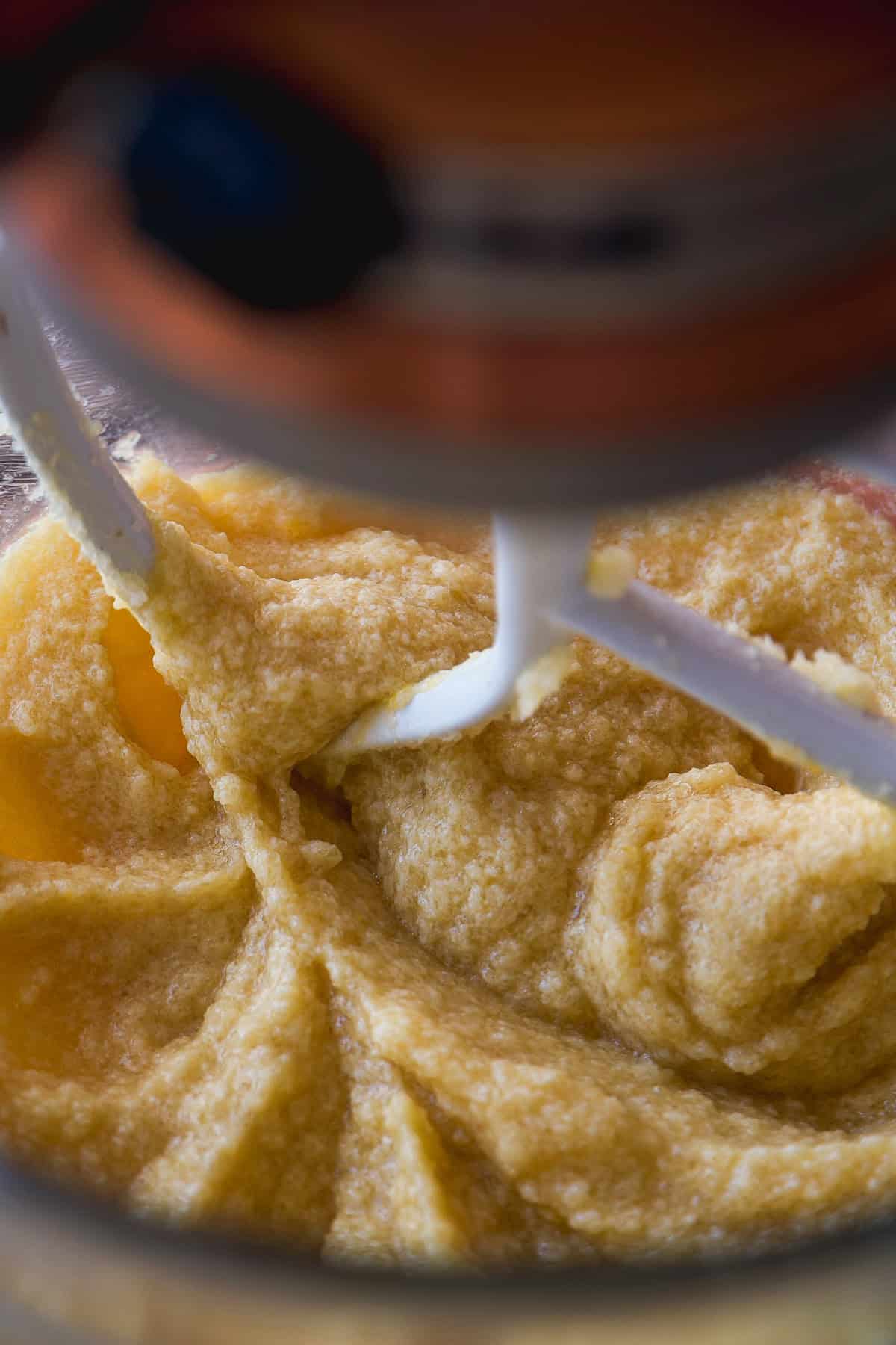 Close-up of thick, yellowish batter being mixed by a stand mixer with a paddle attachment. The texture is slightly grainy, and the mixer bowl and part of the mixer are visible.