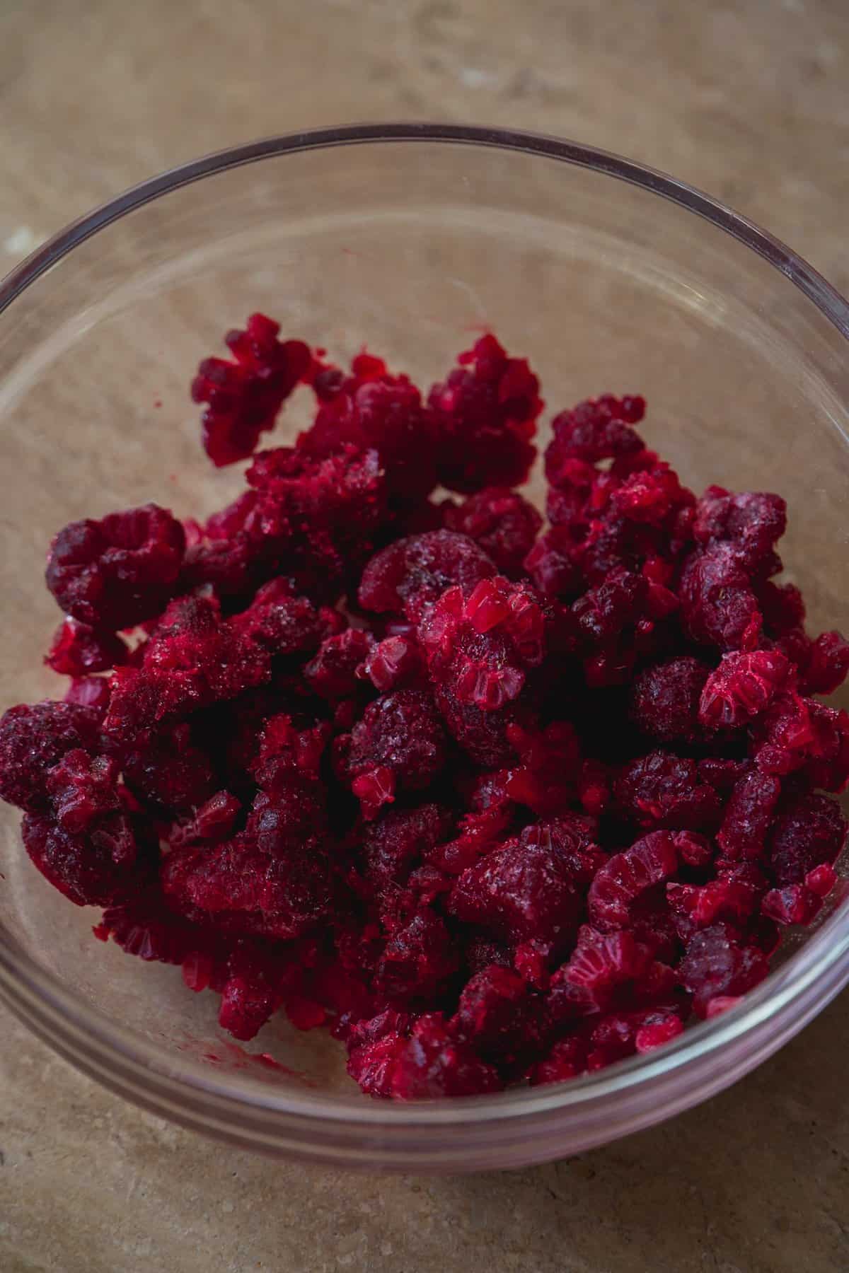 A clear glass bowl filled with frozen raspberries sits on a beige surface. The raspberries are bright red and covered with a light layer of frost.