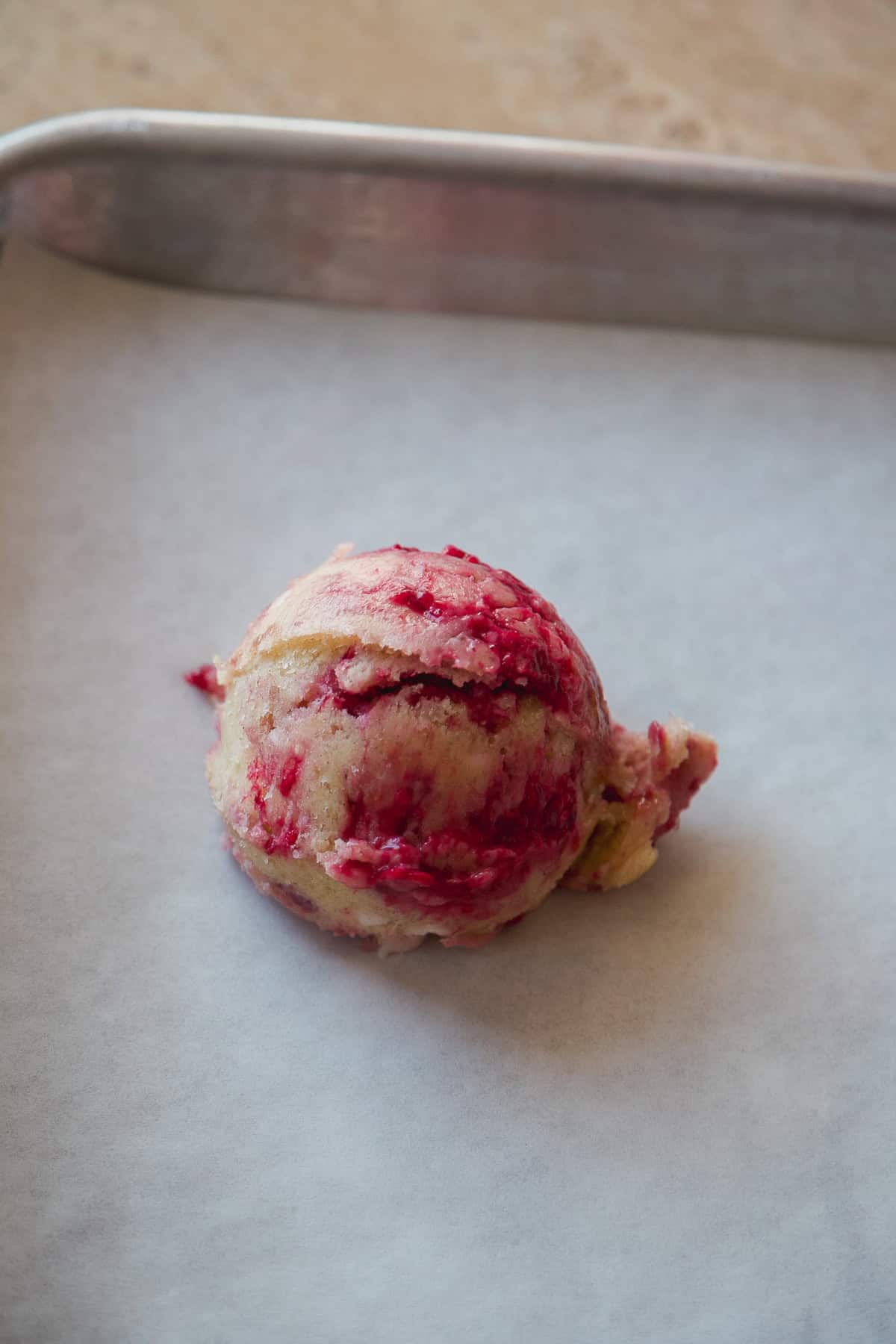 A scoop of cookie dough with red and yellow swirls sits on a parchment-lined baking sheet.