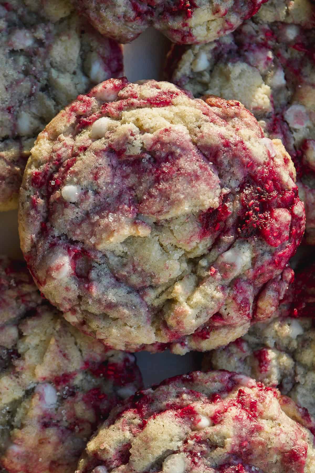 Close-up of cookies with a marbled appearance, featuring white chocolate chips and vibrant red raspberry swirls throughout the golden-brown dough.