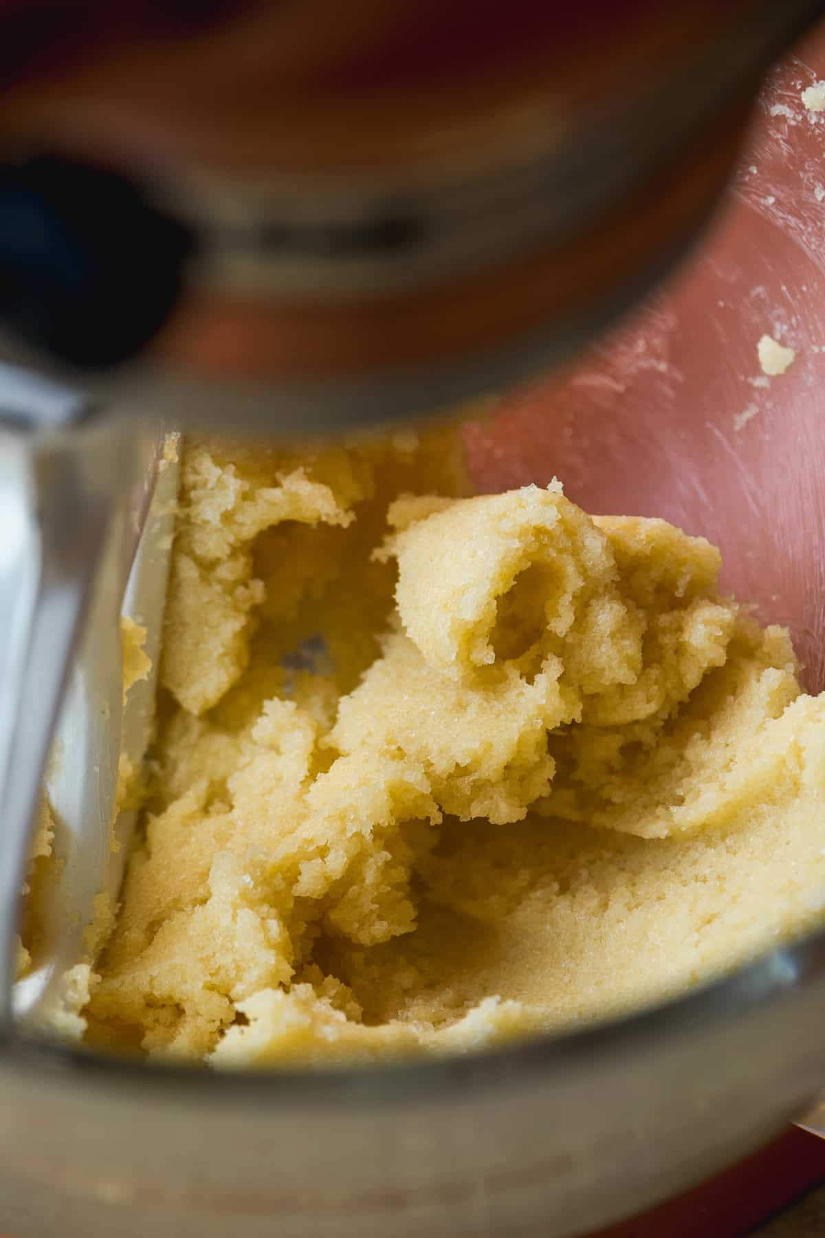 A close-up view of cookie dough being mixed in a stand mixer with a metal paddle attachment. The dough is creamy and light yellow, and the mixing bowl is partially visible.