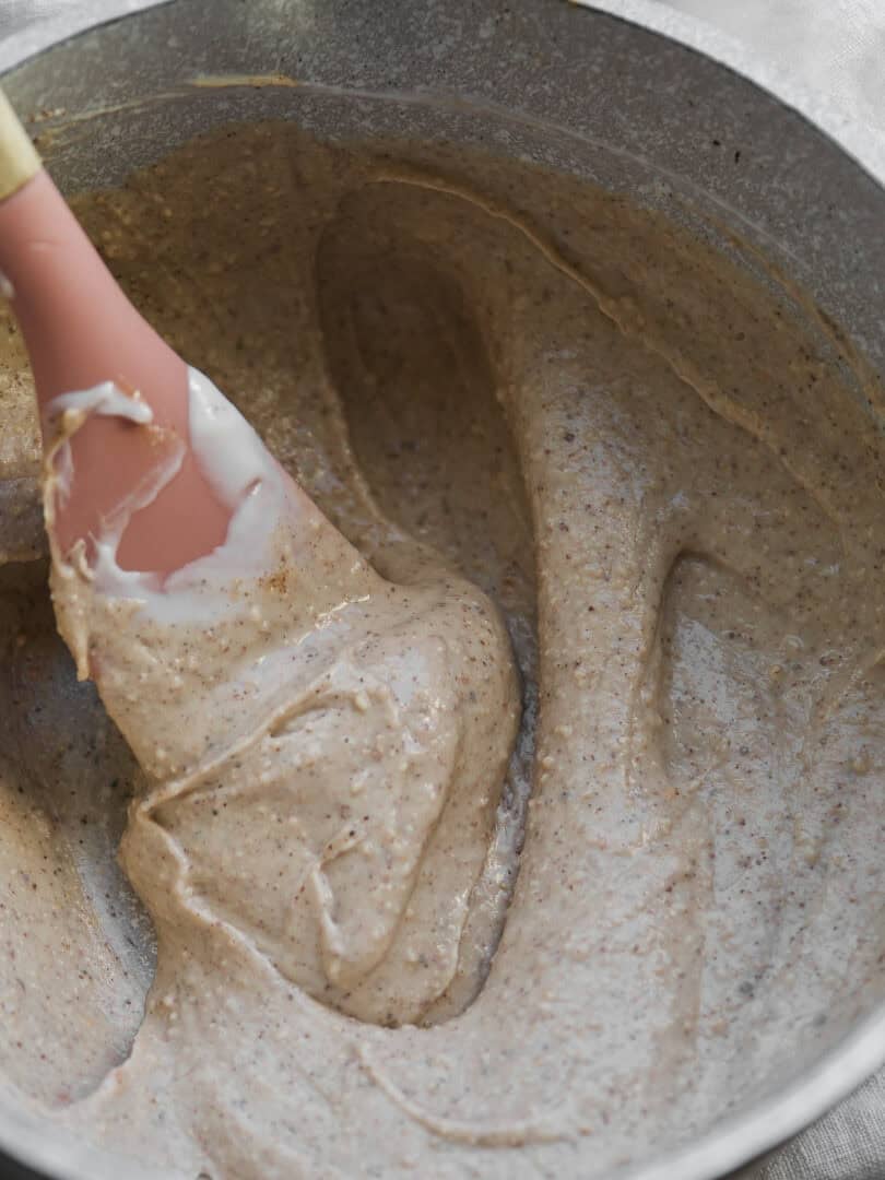 A close-up of a mixing bowl with a thick, speckled batter being stirred by a pink spatula. The batter appears to be partially mixed, with some streaks visible.