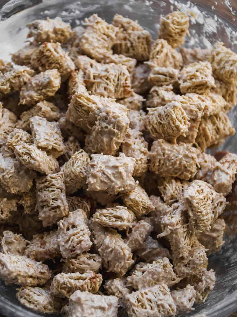A close-up of a glass bowl filled with cereal squares coated in a creamy, white mixture, resembling a dessert or snack preparation.