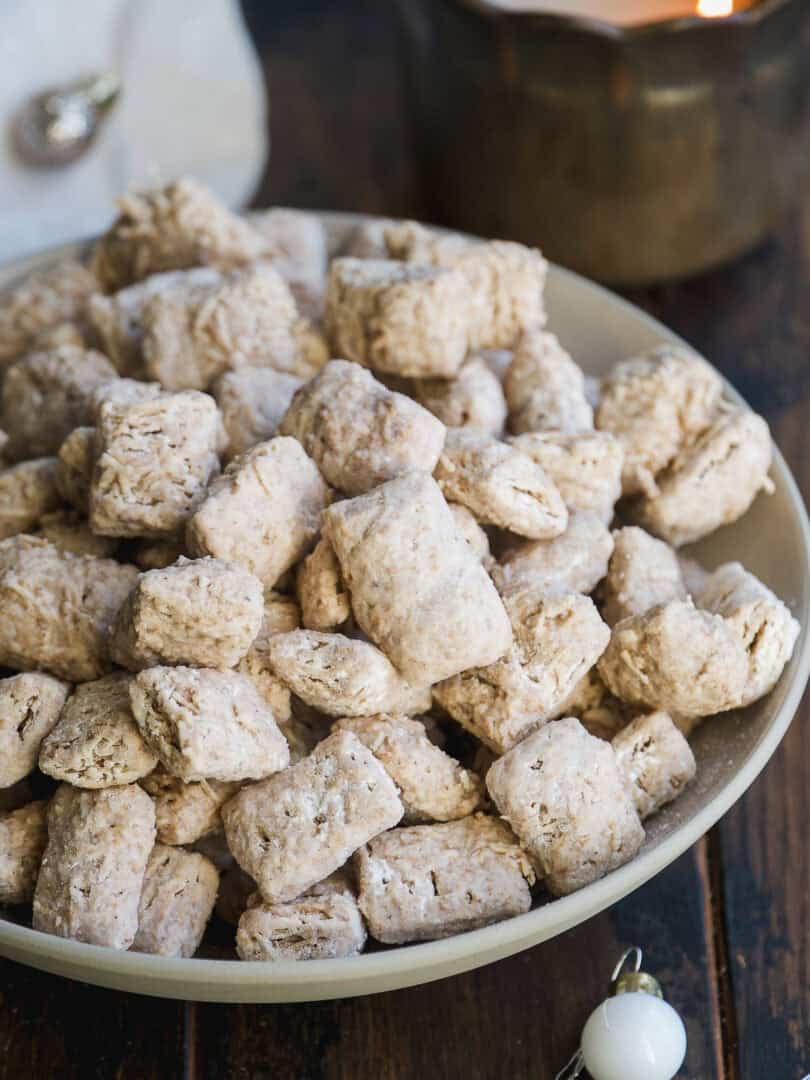 A bowl filled with powdered sugar-coated bite-sized cereal pieces sits on a wooden table, with a lit candle and a white ornament in the background.