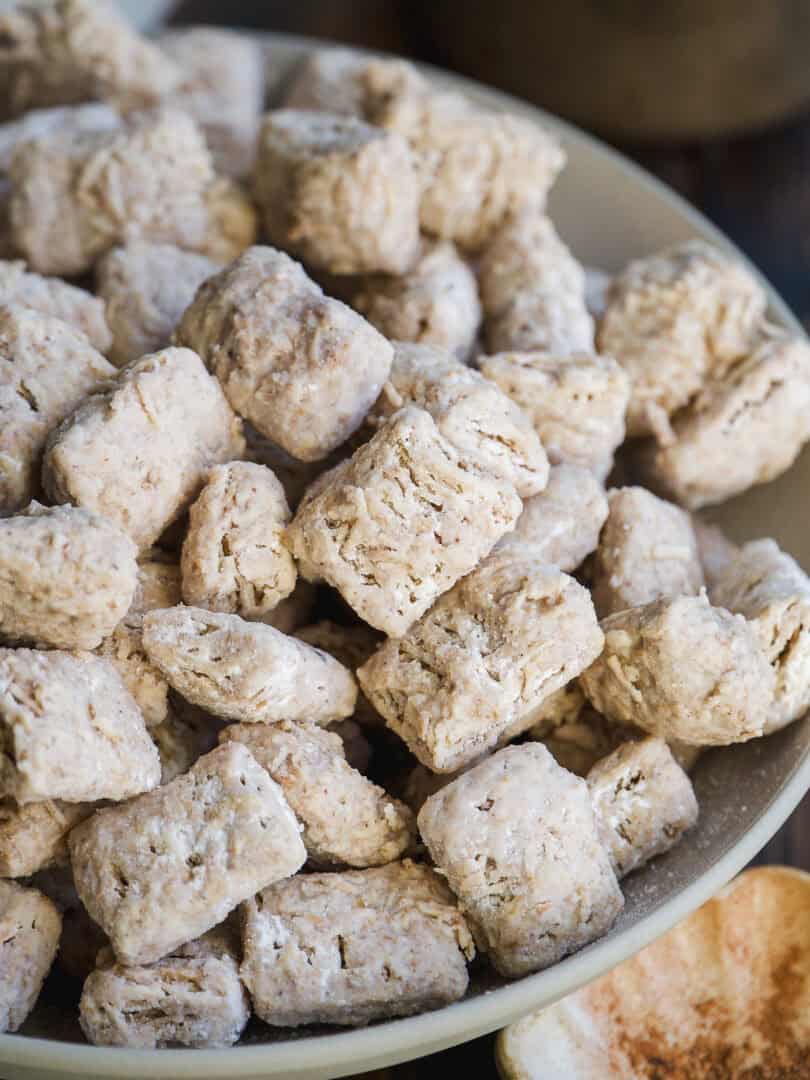 A close-up of a bowl filled with square, wheat cereal biscuits, showing their textured, lightly powdered surfaces. Nearby, a small dish contains ground cinnamon or brown sugar.