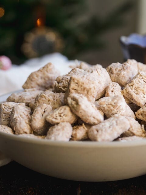 A close-up of a bowl filled with powdered sugar-coated cereal treats, also known as puppy chow or muddy buddies, on a wooden surface with a blurred background.