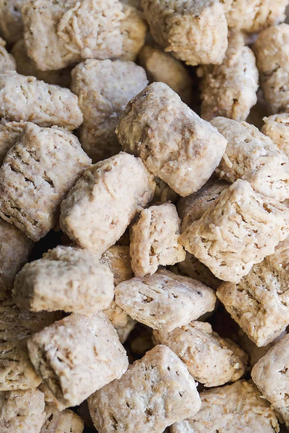 Close-up of a pile of bite-sized shredded wheat cereal pieces, featuring a woven, textured surface and a light brown color.