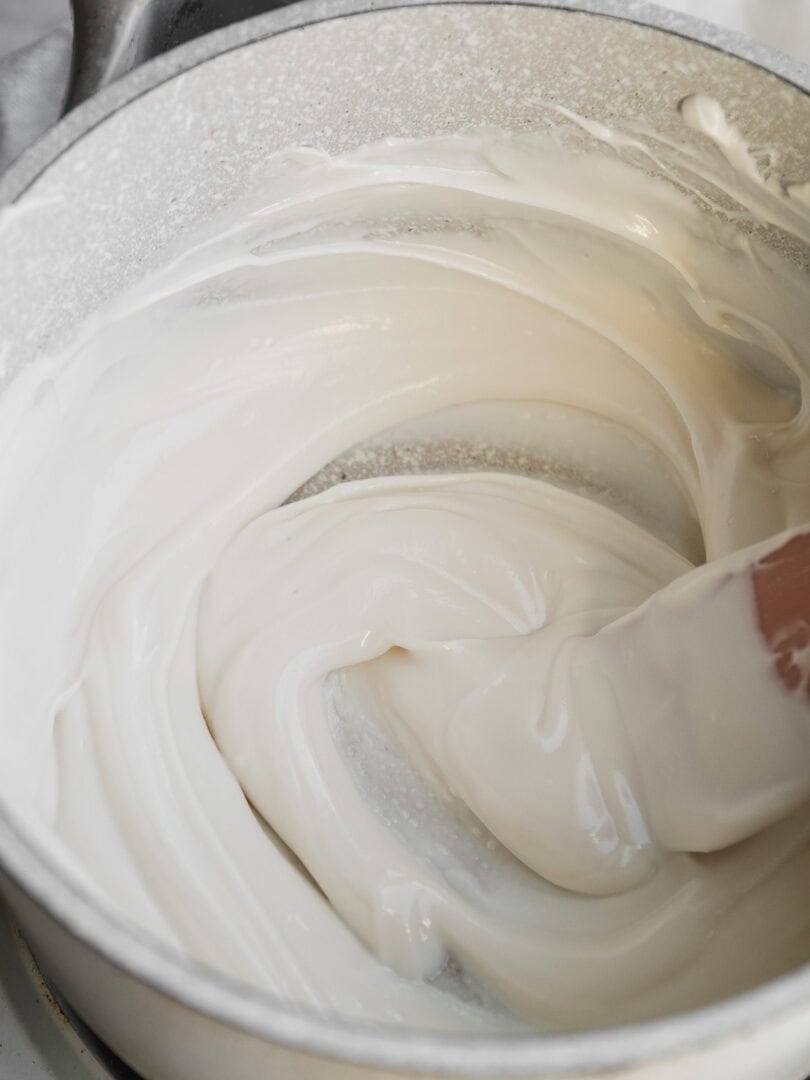 A close-up of a white saucepan containing melted white chocolate or a creamy mixture being stirred with a spatula.