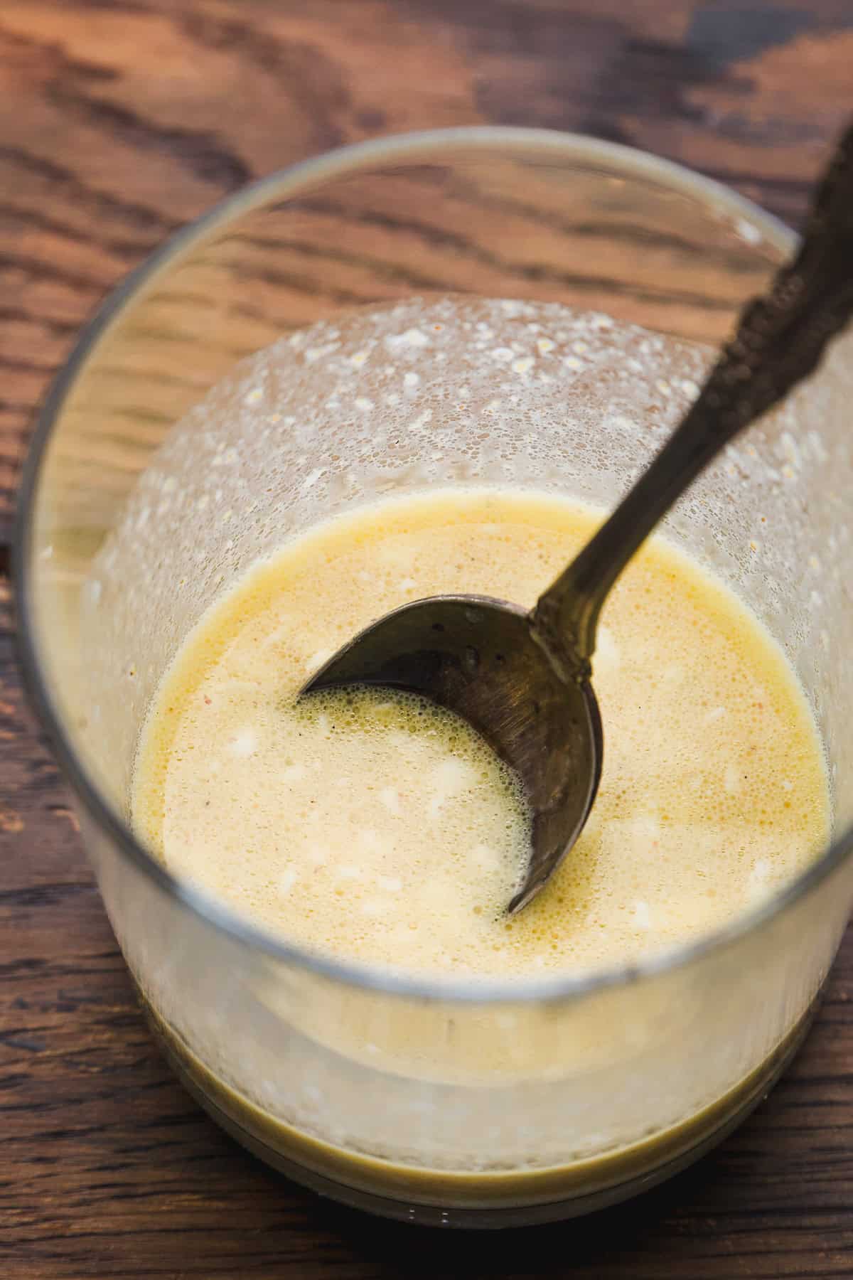 A glass bowl with a creamy yellow liquid mixture and a metal spoon, sitting on a wooden surface. The mixture appears to have small white bits, suggesting its a dressing or sauce being prepared.
