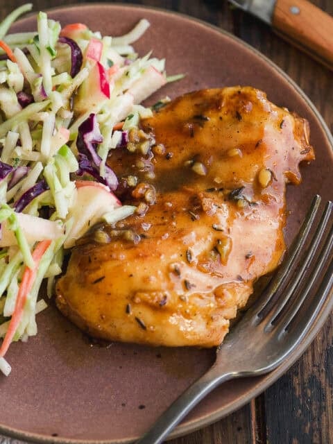 A plate with glazed baked chicken breast next to a fresh cabbage and apple slaw, with a fork resting on the plate and a wooden-handled knife in the background.