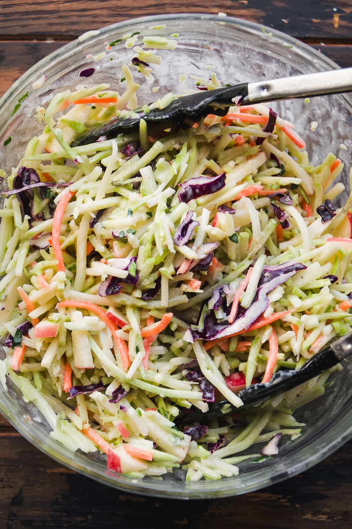 A glass bowl filled with mixed coleslaw containing shredded green cabbage, purple cabbage, and carrots, with a creamy dressing. Black tongs rest on the bowl’s edge.