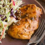 A plate with a glazed, seasoned chicken breast and a side of colorful cabbage slaw, with a fork next to the food on a rustic wooden table.