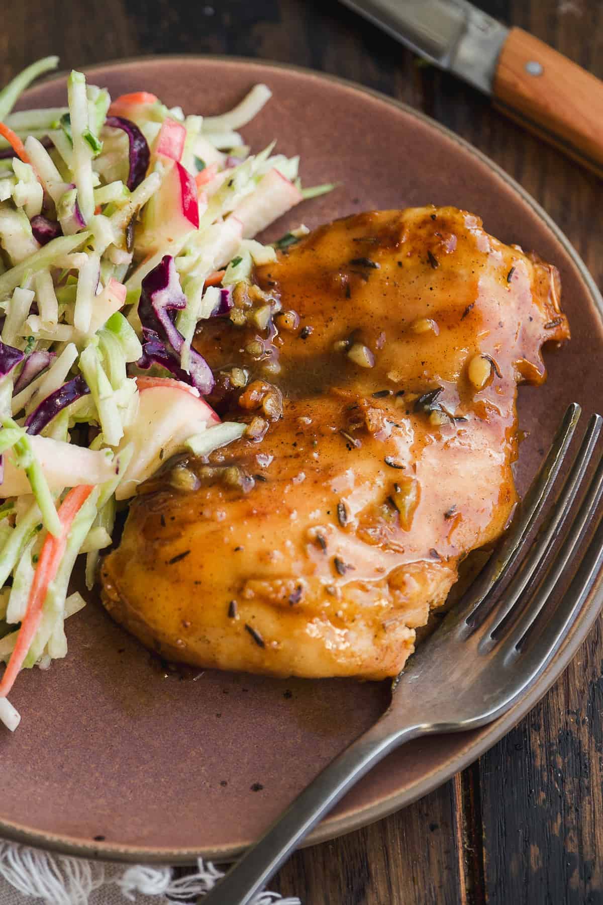 A plate with glazed baked chicken breast and a side of colorful cabbage slaw with carrots and apple slices. A fork rests beside the chicken, and a knife is partially visible in the background.