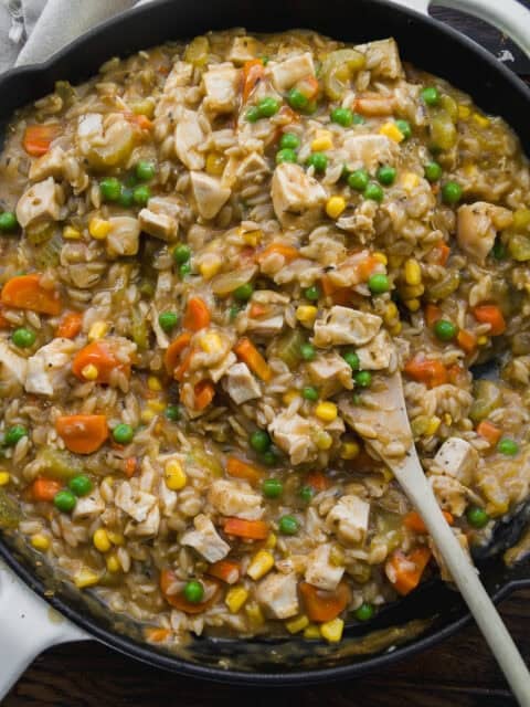 A skillet filled with a creamy mixture of chopped chicken, peas, corn, carrots, celery, and rice, being stirred with a wooden spoon. The dish looks hearty and colorful.