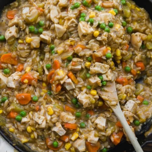 A skillet filled with a creamy mixture of chopped chicken, peas, corn, carrots, celery, and rice, being stirred with a wooden spoon. The dish looks hearty and colorful.