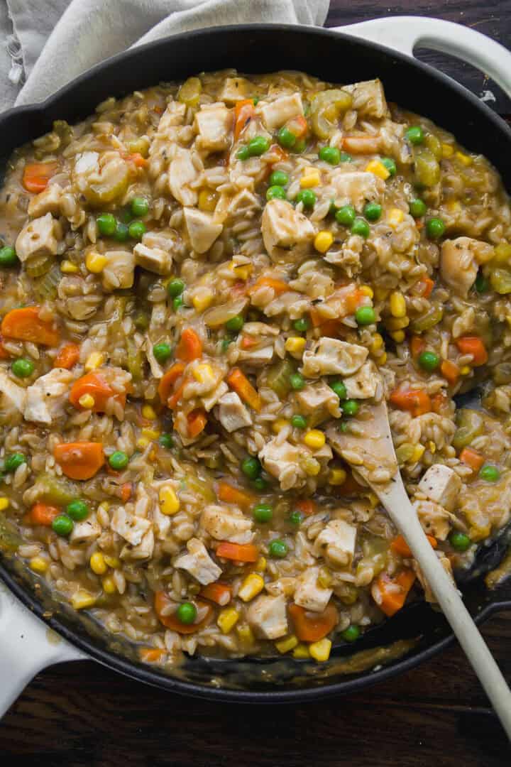 A skillet filled with a creamy mixture of chopped chicken, peas, corn, carrots, celery, and rice, being stirred with a wooden spoon. The dish looks hearty and colorful.
