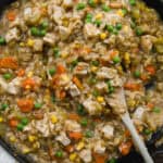 A skillet filled with creamy chicken and rice mixed with peas, carrots, corn, and celery. A wooden spoon rests in the dish, ready for serving.