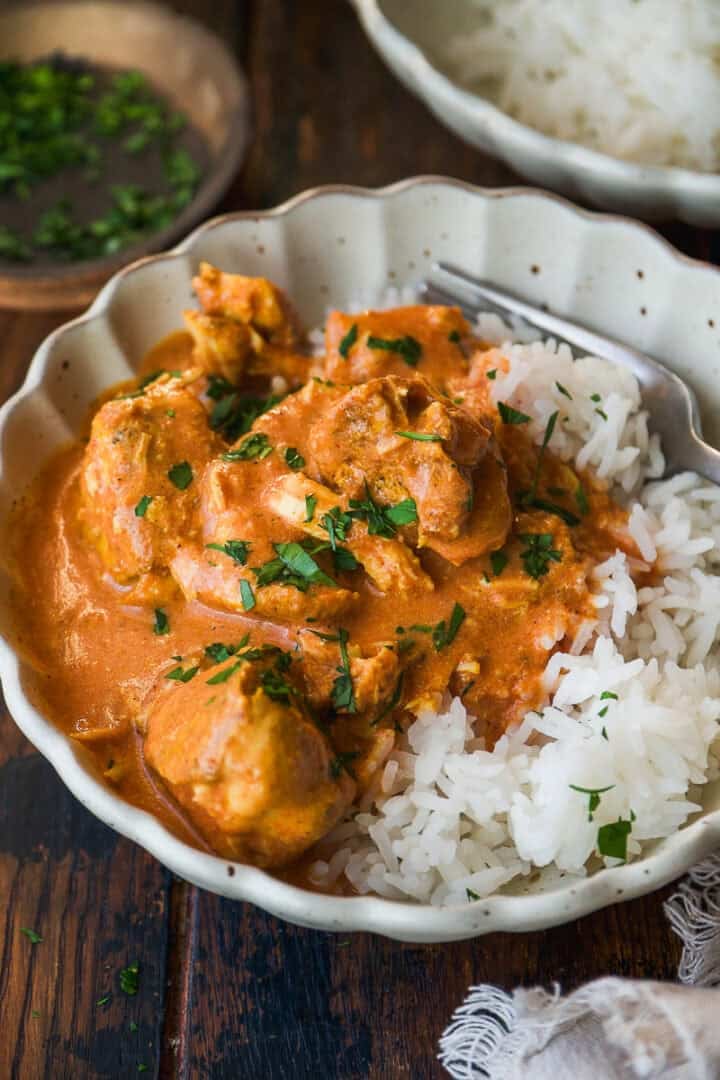 A bowl of white rice topped with creamy orange chicken curry, garnished with chopped fresh herbs, sits on a rustic wooden table.
