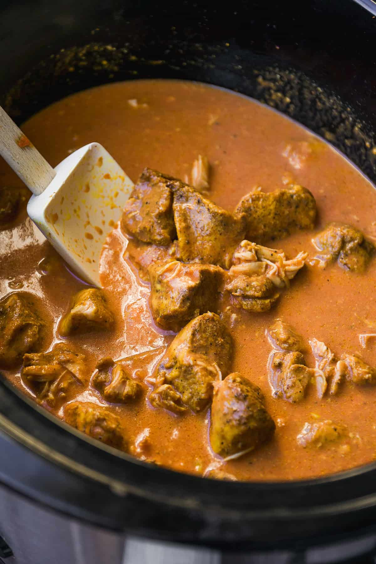 Chunks of cooked chicken in a rich, orange-red curry sauce inside a slow cooker, being stirred with a white spatula.