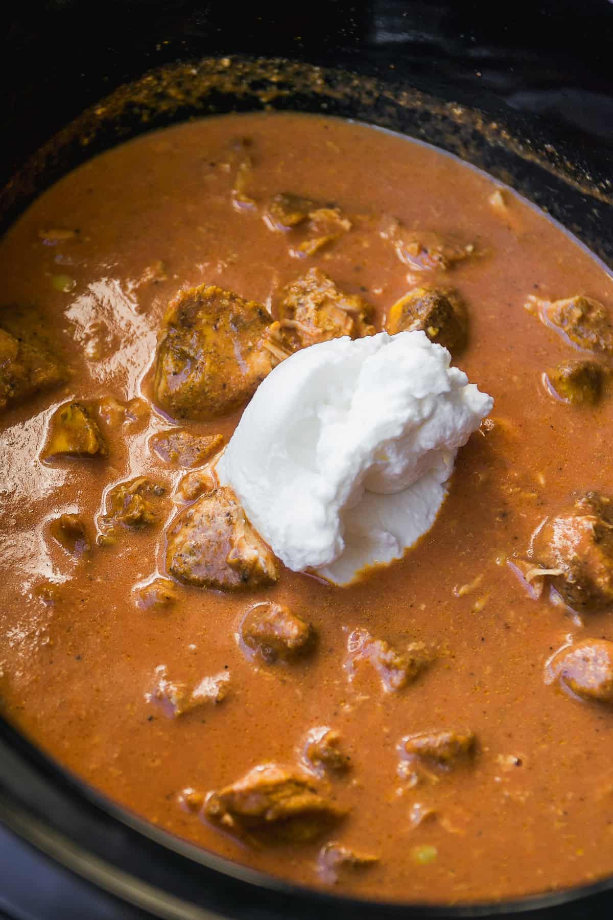 Chunks of meat in a rich, reddish-brown sauce simmering in a pot, topped with a dollop of white cream or yogurt in the center.