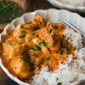 A bowl of white rice topped with creamy orange chicken curry, garnished with fresh chopped cilantro, with a fork resting on the side. Another bowl of rice and a small dish of herbs are in the background.