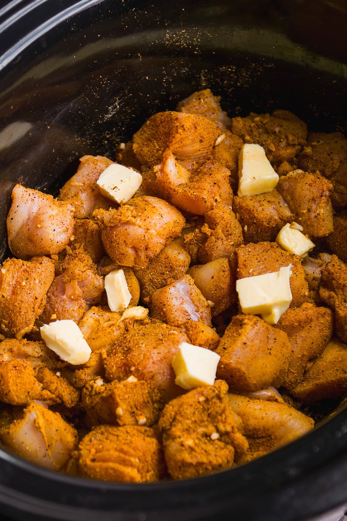 Chunks of raw, seasoned chicken with several small cubes of butter are placed inside a black slow cooker, ready to be cooked.