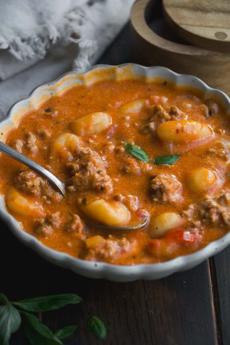 A white bowl filled with creamy tomato soup containing gnocchi and ground meat, garnished with fresh basil. A spoon rests in the bowl, and a wooden container and napkin are visible in the background.