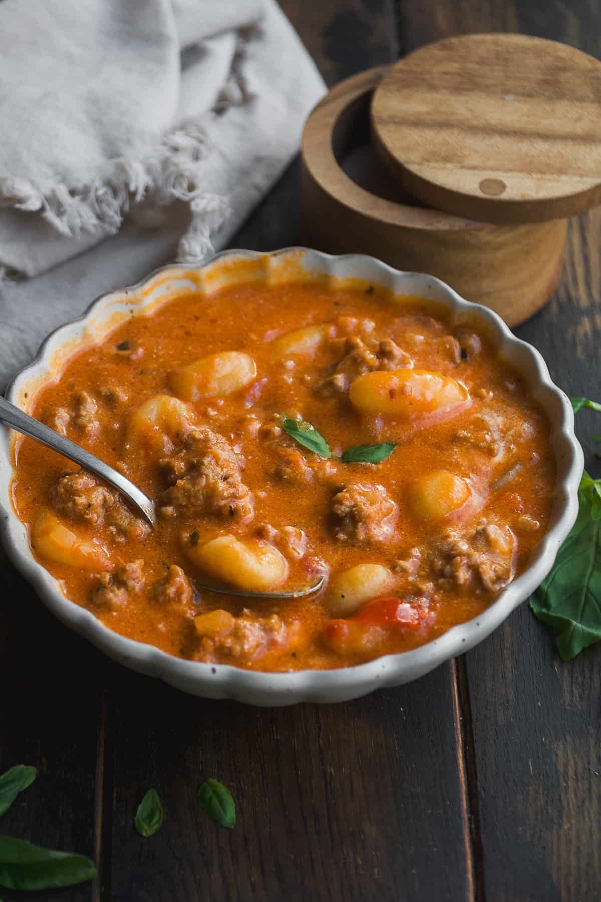 A bowl of creamy tomato soup with gnocchi, ground meat, and fresh basil, served with a spoon on a wooden table. A cloth napkin and a wooden container are in the background.