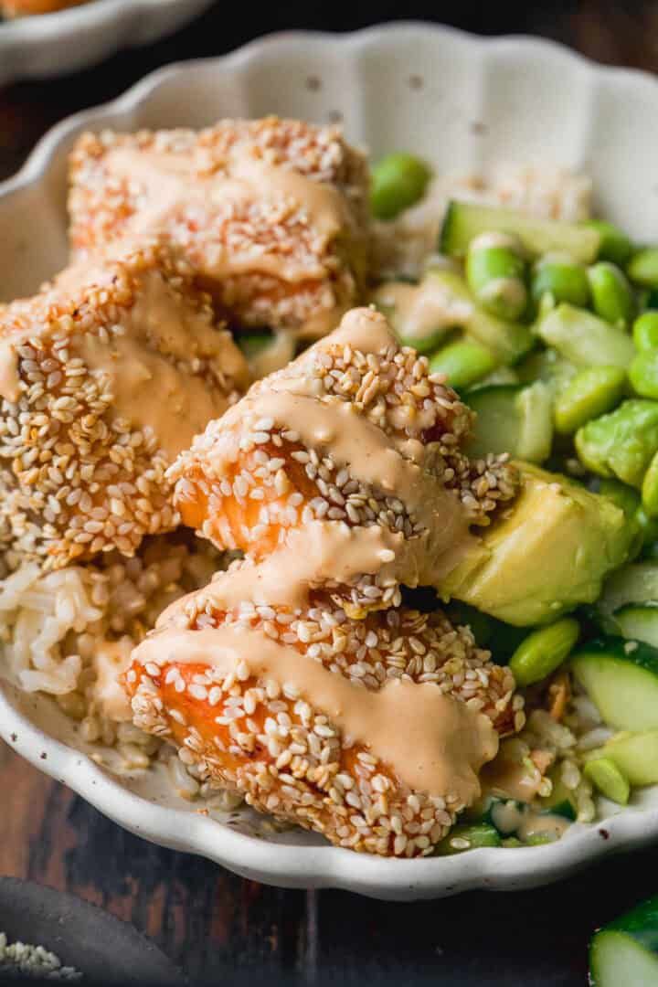 A bowl with sesame-crusted salmon pieces drizzled with creamy sauce, served over rice with sliced avocado and edamame on the side.