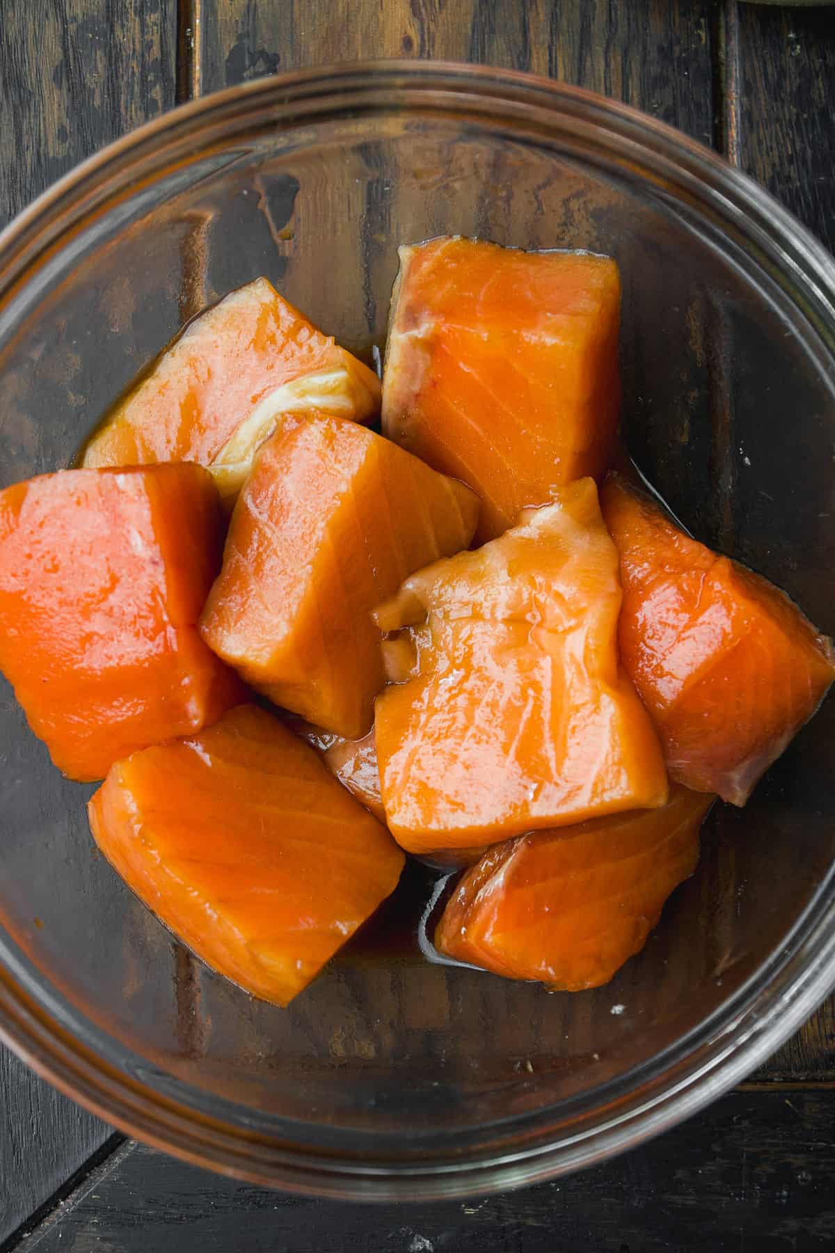 Several chunks of raw, orange-colored salmon are placed in a clear glass bowl, set on a dark wooden surface. The fish pieces appear fresh and glossy, ready for cooking or marinating.