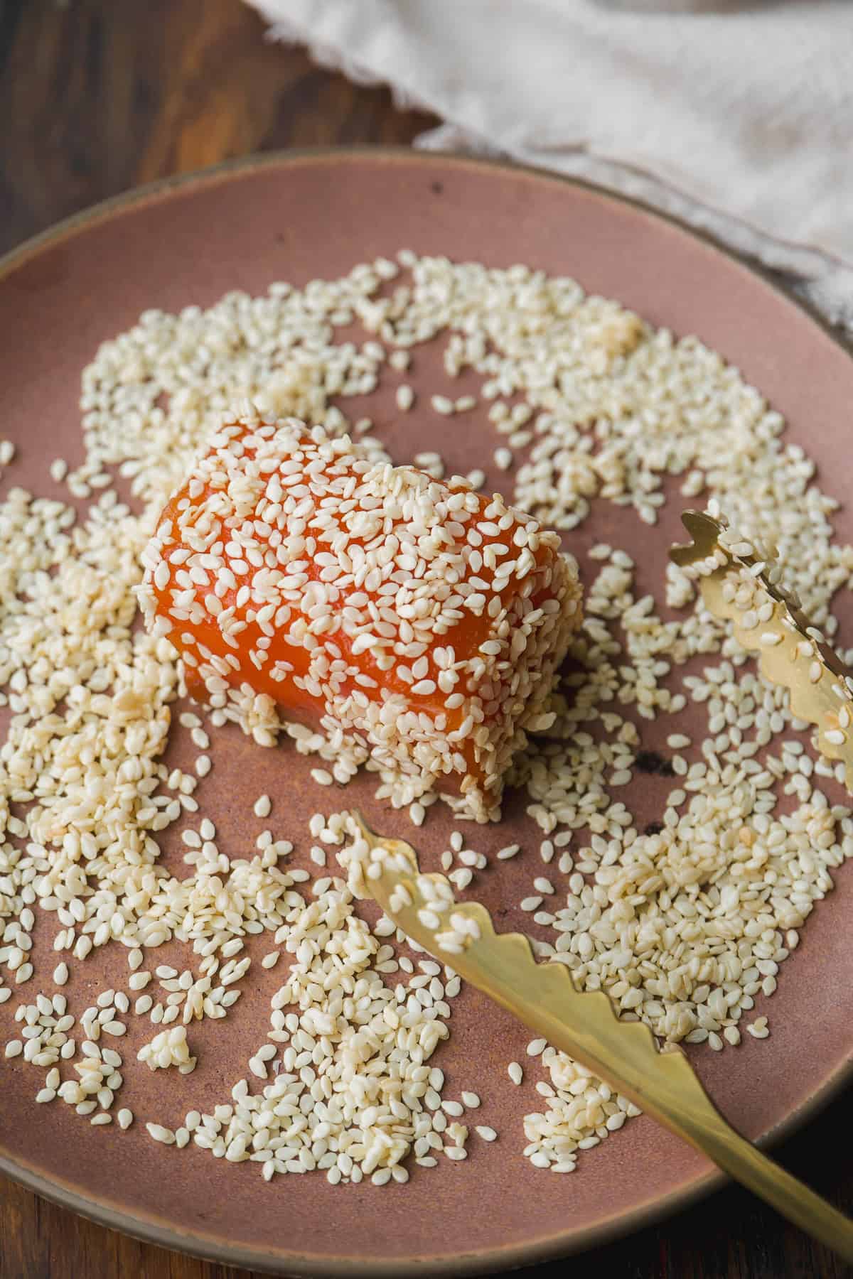 A piece of orange-colored food, possibly cheese or tofu, is coated in sesame seeds on a brown plate. Golden tongs rest nearby, and sesame seeds are scattered around on the plate.