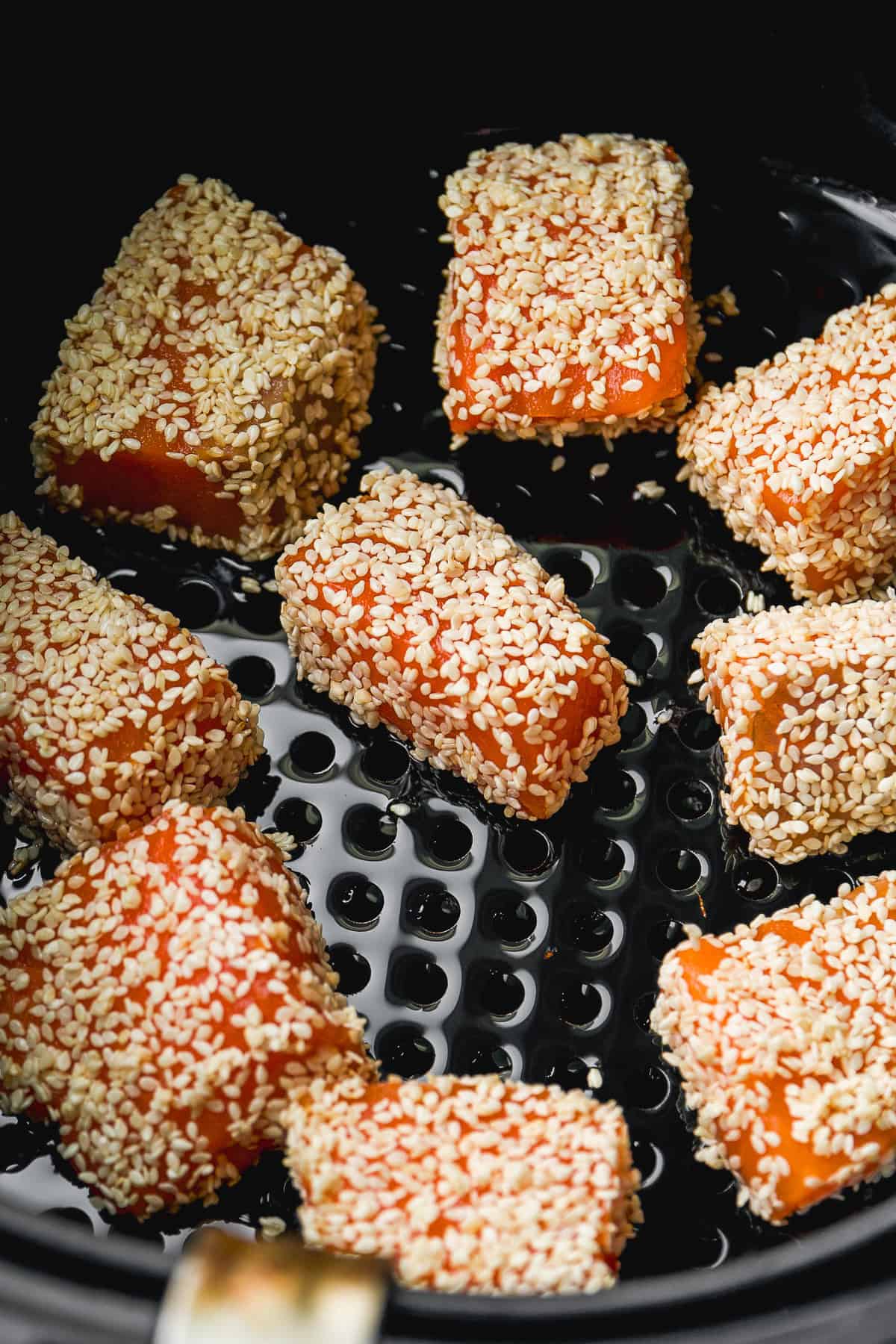 Several pieces of food, likely salmon, coated in sesame seeds are arranged inside an air fryer basket, ready to be cooked.
