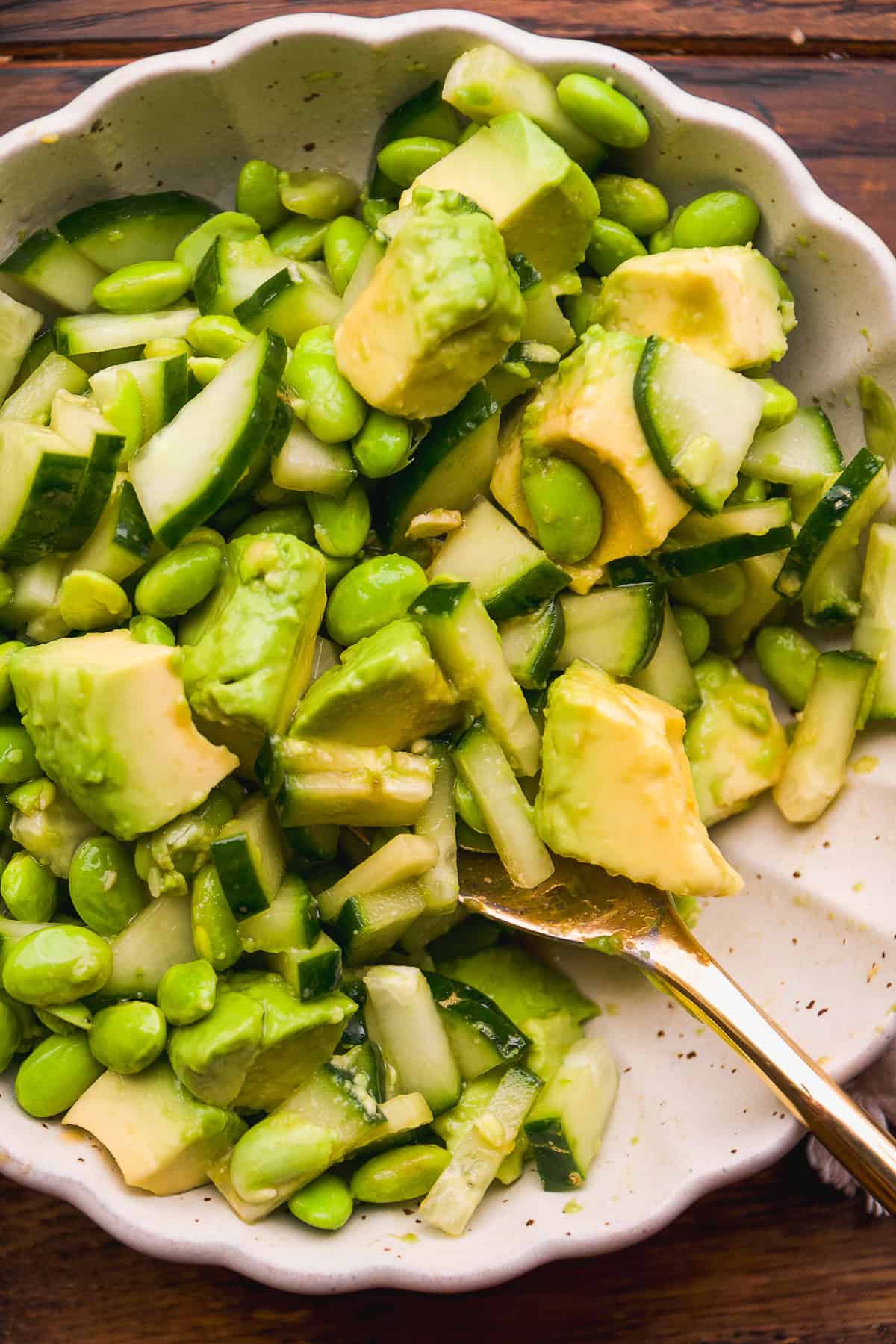 A bowl of fresh salad containing chunks of avocado, sliced cucumber, edamame beans, and possibly onions, with a gold-colored fork resting in the bowl. The salad appears vibrant and healthy.