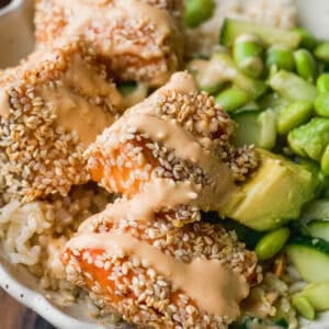 A close-up of a bowl containing sesame-crusted salmon pieces drizzled with a creamy sauce, served with sliced avocado, edamame, cucumber, and rice.