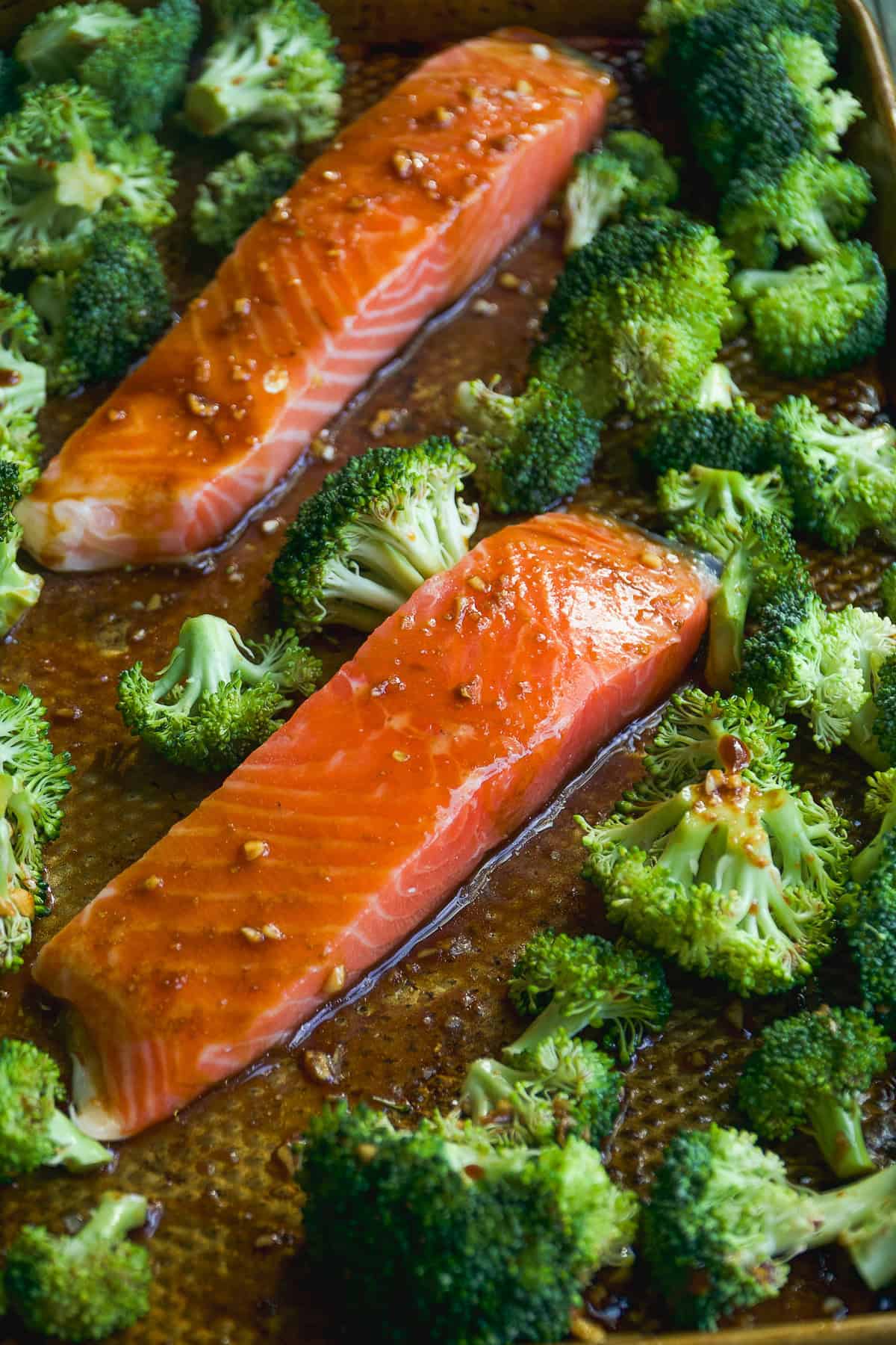 Two glazed salmon fillets and fresh broccoli florets on a baking sheet, ready for roasting. The salmon has a shiny, seasoned surface, surrounded by vibrant green broccoli pieces.