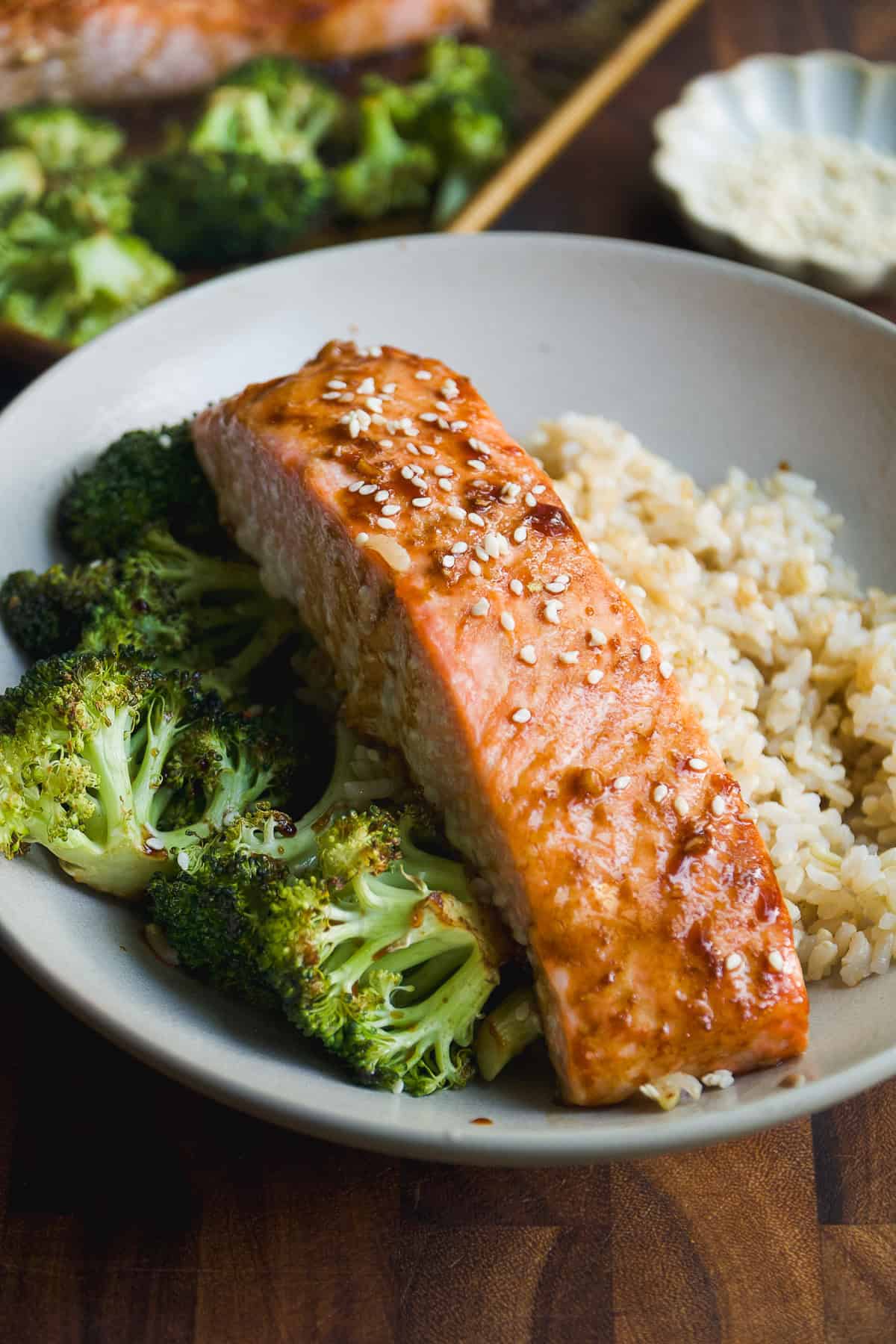 A plate with a glazed salmon fillet topped with sesame seeds, served alongside roasted broccoli and a portion of brown rice.