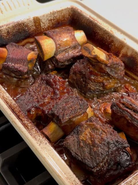 A rectangular baking dish filled with braised beef short ribs in a rich, dark sauce, fresh from the oven and resting on a stovetop. The meat looks tender and browned with visible bones.