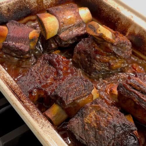 A rectangular baking dish filled with braised beef short ribs in a rich, dark sauce, fresh from the oven and resting on a stovetop. The meat looks tender and browned with visible bones.
