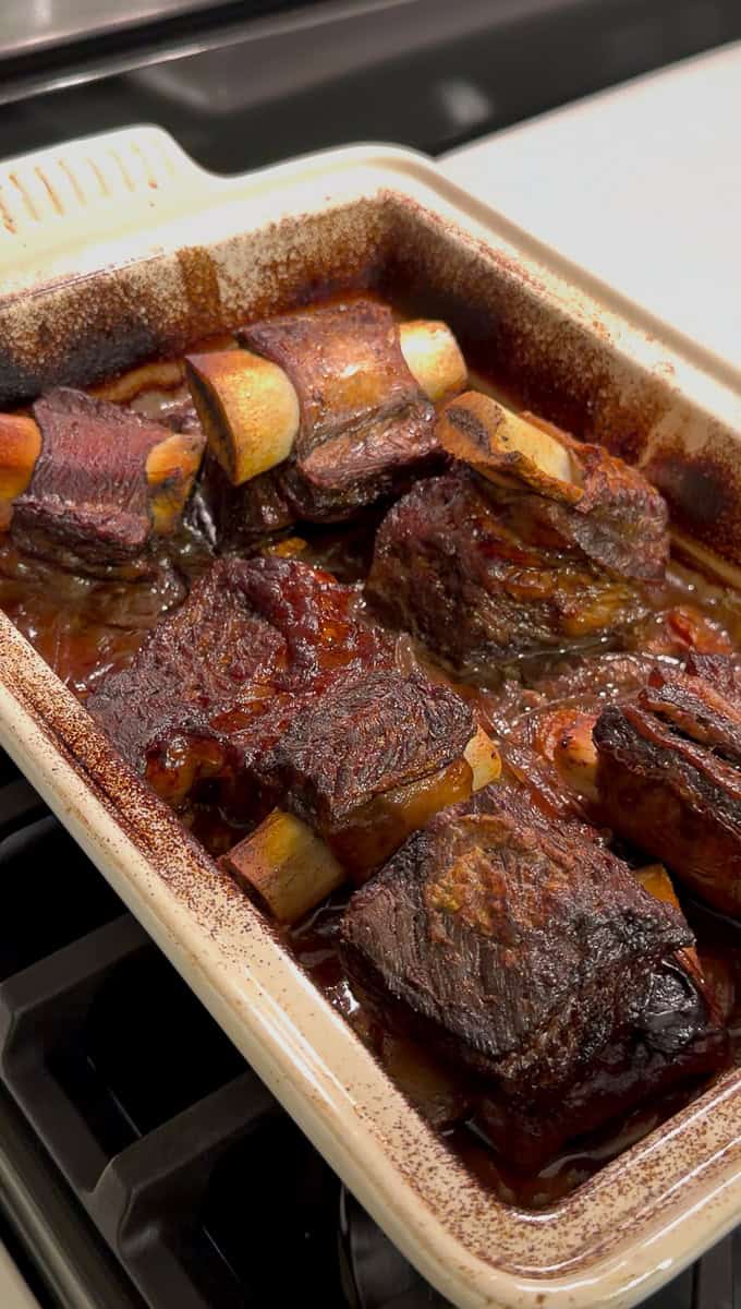 A rectangular baking dish filled with braised beef short ribs in a rich, dark sauce, fresh from the oven and resting on a stovetop. The meat looks tender and browned with visible bones.