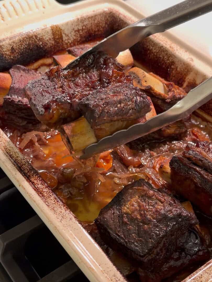 A baking dish filled with braised beef short ribs in a rich, brown sauce with onions. Metal tongs are lifting one rib, showing the caramelized, tender meat and bones. The edges of the dish have baked-on sauce.
