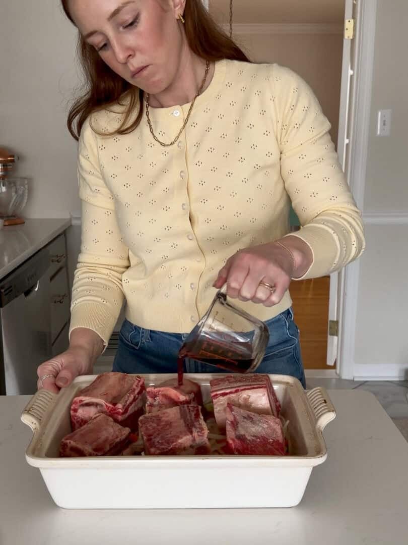 A woman wearing a light yellow sweater pours liquid from a glass measuring cup over raw beef short ribs in a rectangular baking dish on a kitchen counter.