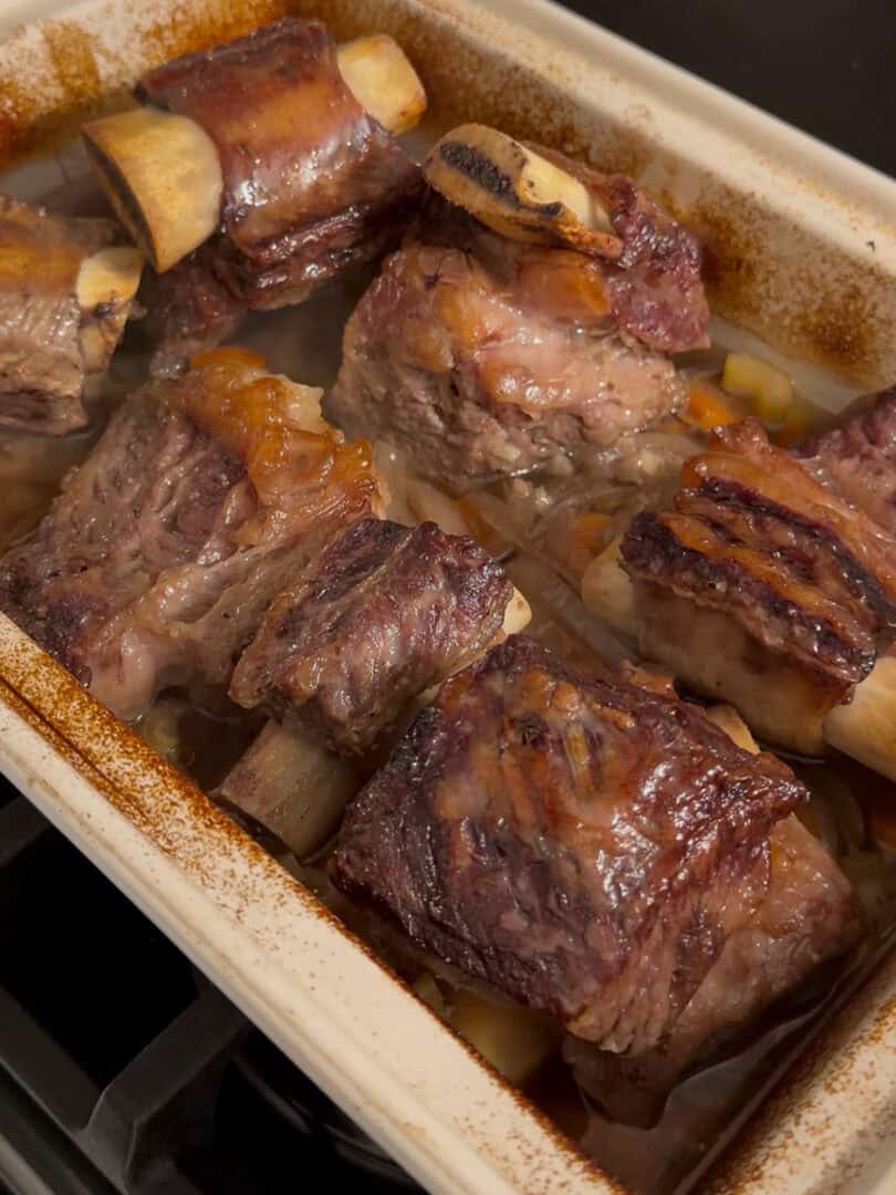 A baking dish filled with braised beef short ribs, featuring browned tops and bones visible, cooking in a savory broth on a stovetop.