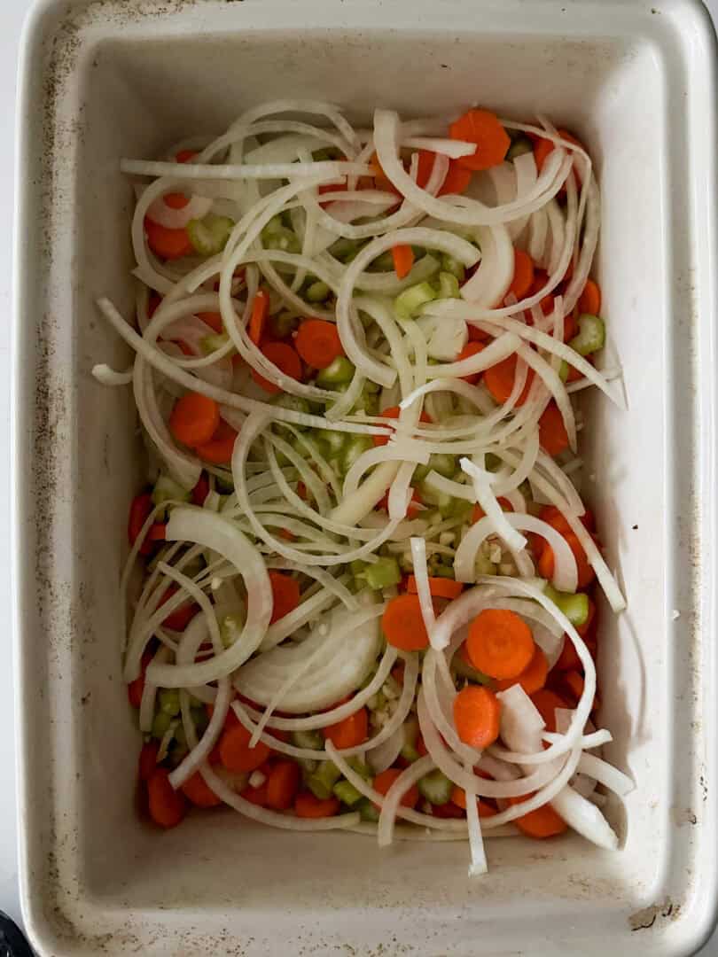 A white baking dish filled with sliced onions, carrots, and celery, arranged in layers and ready for cooking. The vegetables are fresh and colorful, with visible rings of onions and round carrot slices.