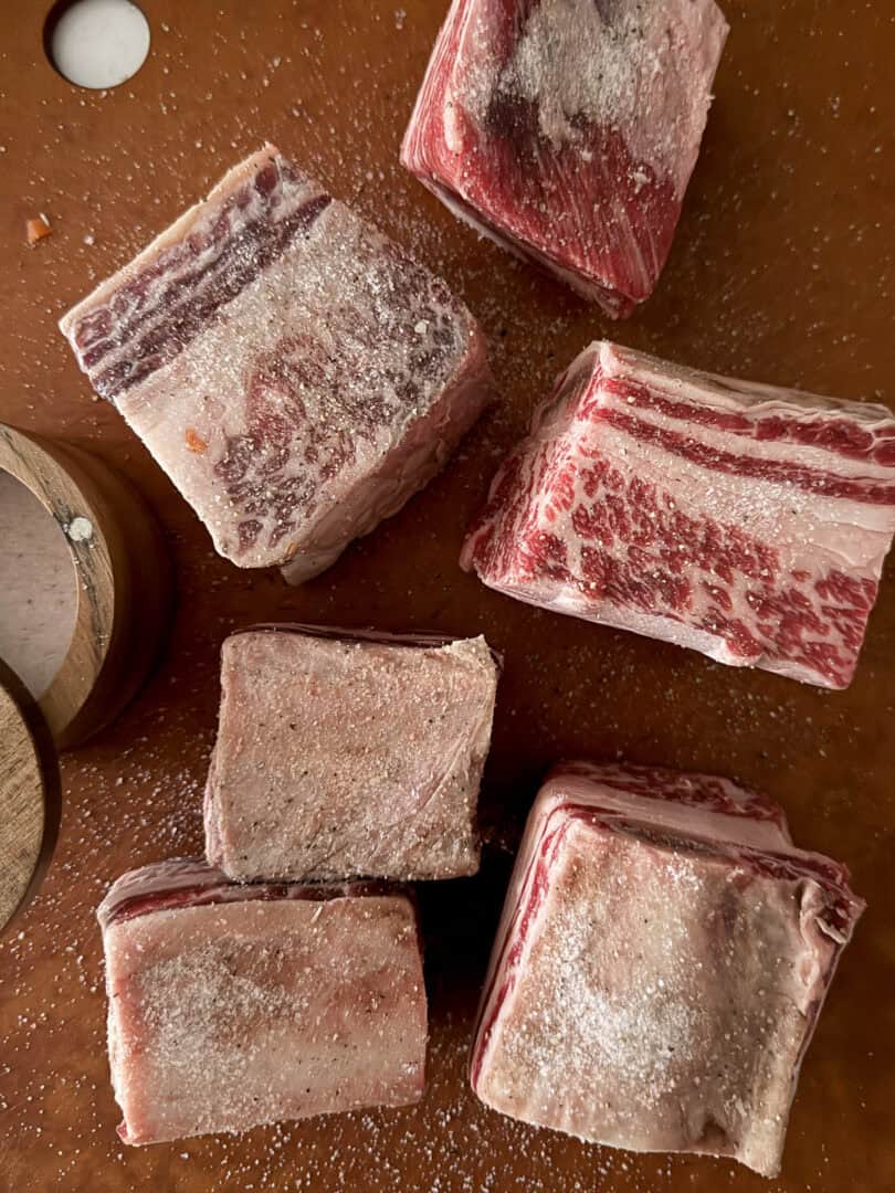 Six raw, marbled beef short ribs are sprinkled with salt on a brown cutting board. A round wooden salt cellar sits nearby, partially open.